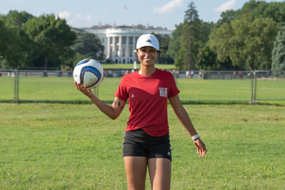 alana pictured in front of white house with soccer ball in red shirt and white hat