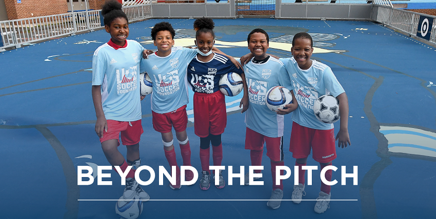 five black children, two girls and three boys, in bright blue soccer jerseys posing for photo on a dark blue mini-pitch