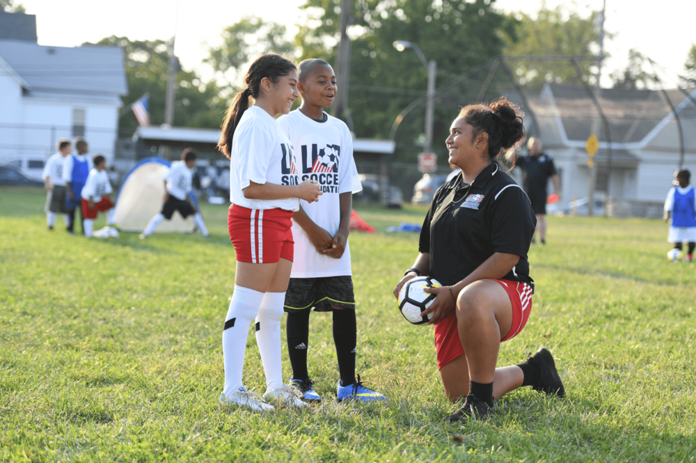 Coach kneeling while holding a soccer ball and talking to a girl and a boy soccer player