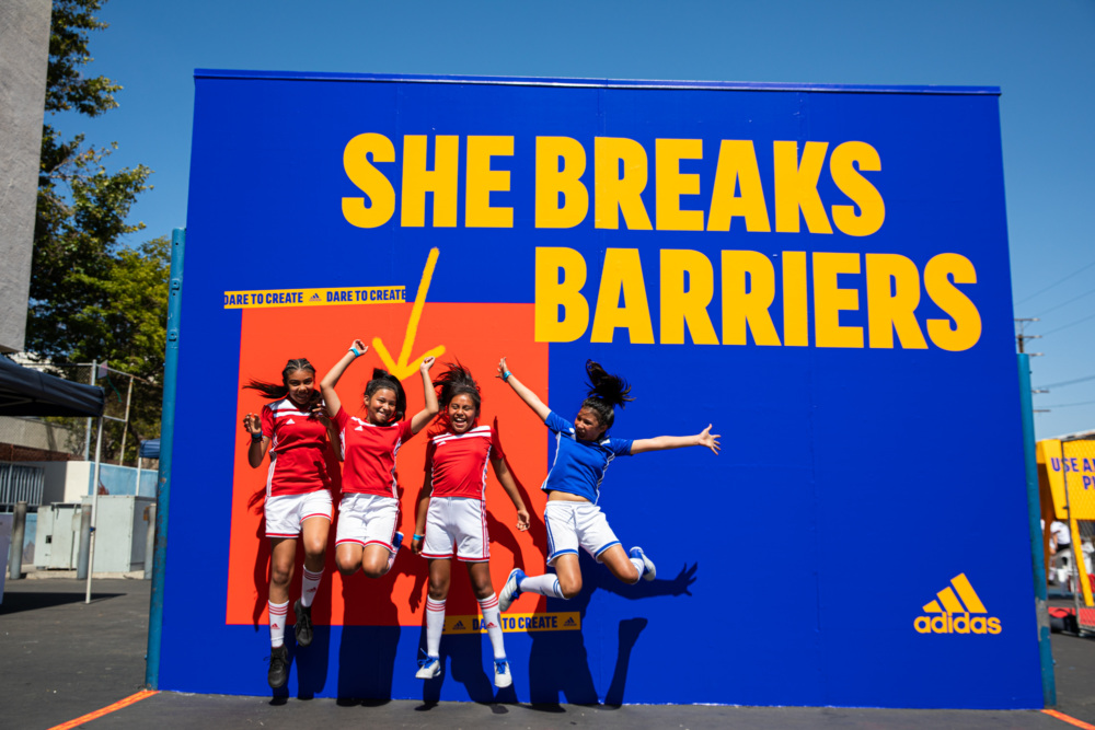 Girls jumping in front of a blue wall that says "She Breaks Barriers"