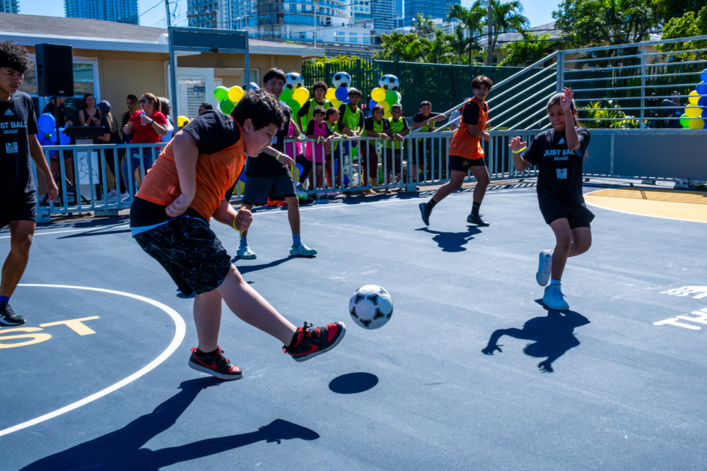 A boy in an orange pinnie kicks a soccer ball on a blue pitch toward a goal just outside of the photo.