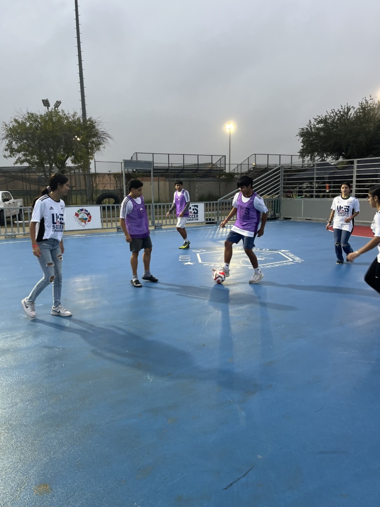 A group of kids half in white shirts and half in purple pinnies play soccer on a blue mini-pitch