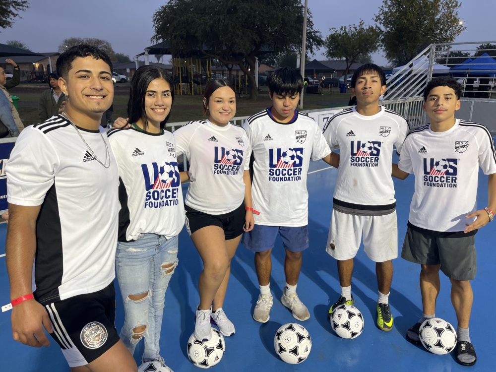 A group of six teenagers stand on a mini-pitch smiling with soccer balls at their feet.
