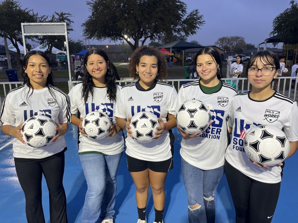 Five girls stand on a blue mini-pitch holding soccer balls