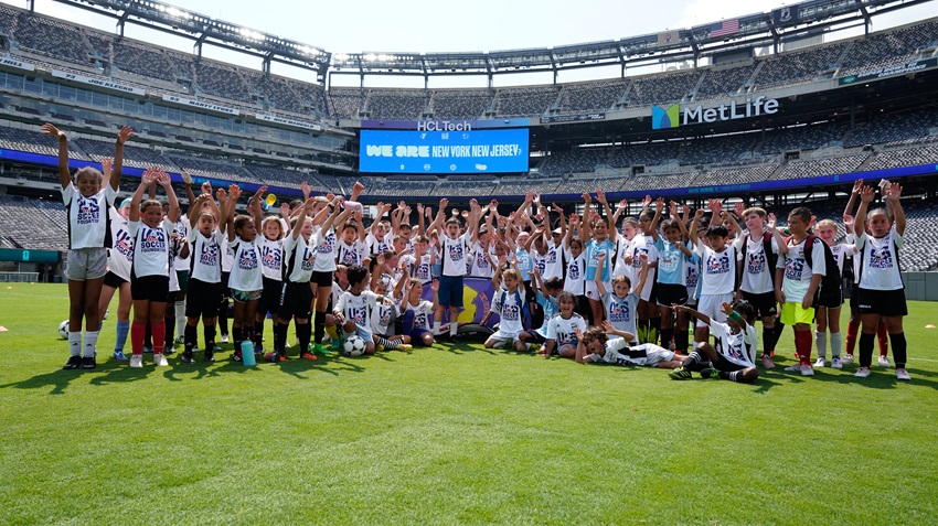 a group of about 60 young kids wearing white U.S. Soccer Foundation jerseys smile at the camera with their hands raised in celebration. they are facing the camera from the field at metlife stadium during a bright, sunny day.