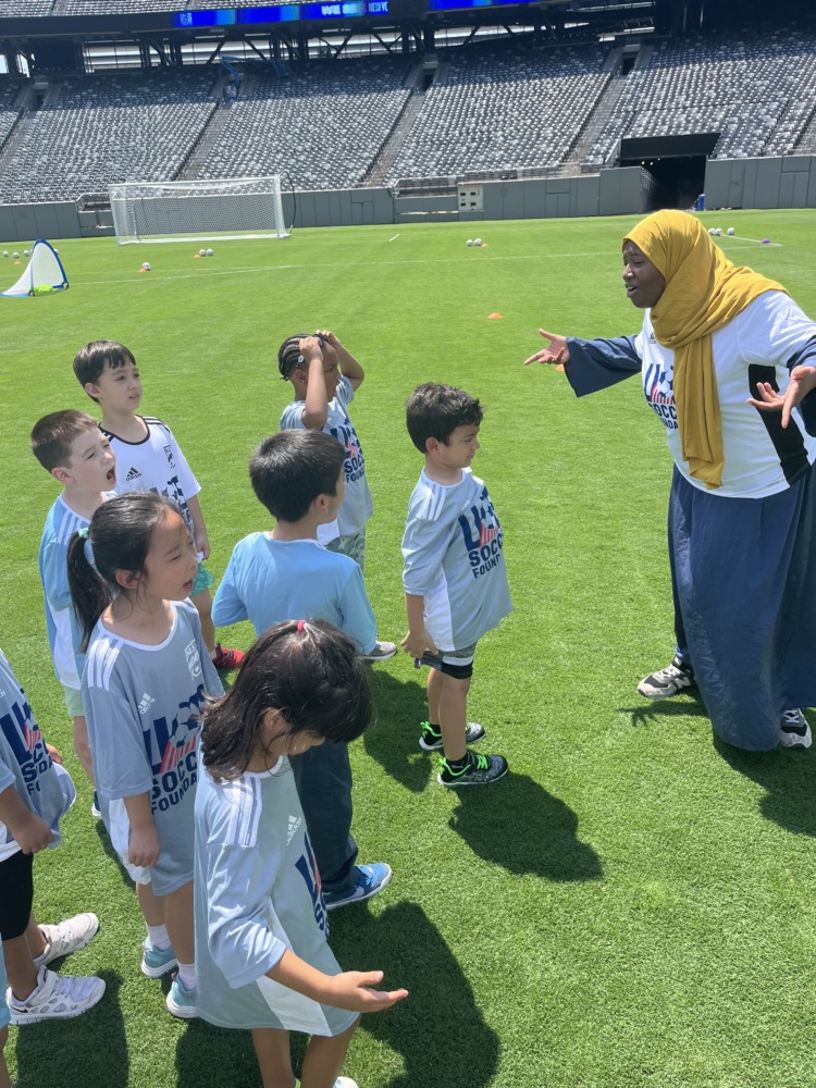 a coach in a yellow hijab, white U.S. Soccer Foundation jersey, and long blue skirt speaks to her team of young kids to get them excited on the MetLife Stadium soccer field. seven young kids wearing light blue U.S. Soccer Foundation jerseys scream back at her in excitement