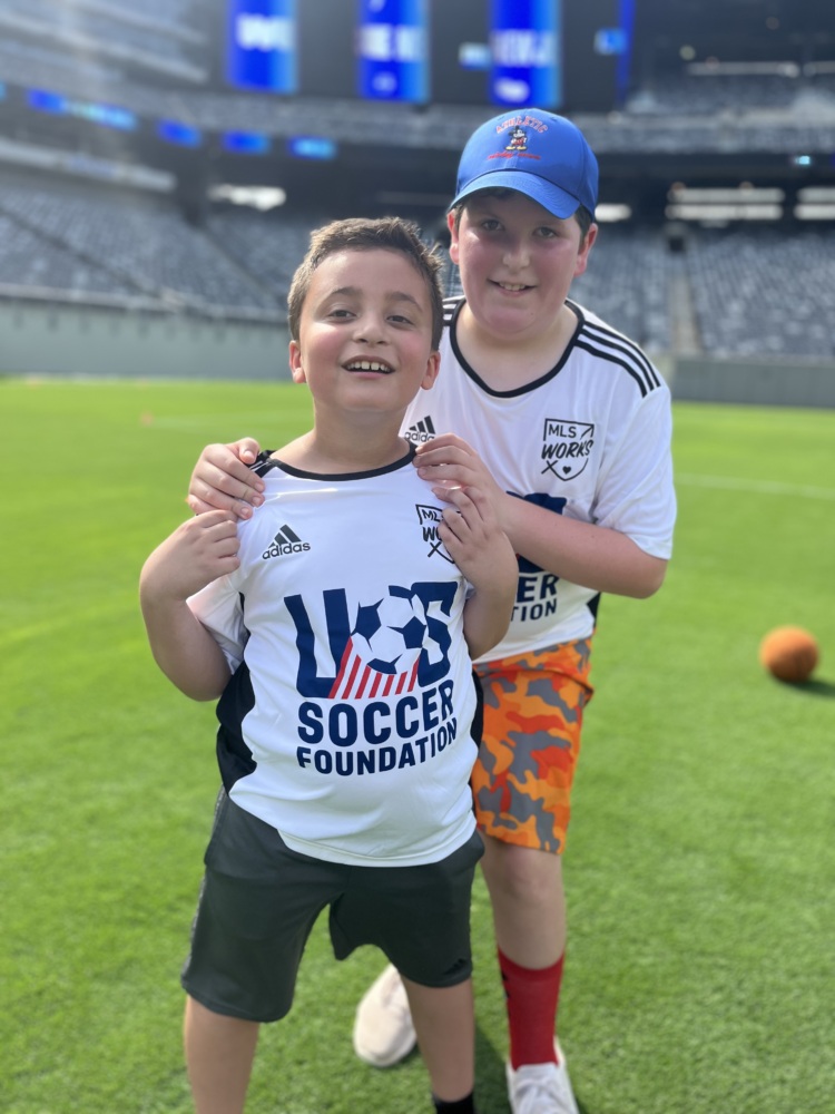 two boys pose together for a photo from the soccer field. they are each wearing a white U.S. Soccer Foundation jersey. the boy in the front has darker hair and is shorter than his friend who is holding his shoulders from just behind him. the other boy is a bit taller and is wearing a blue cap.