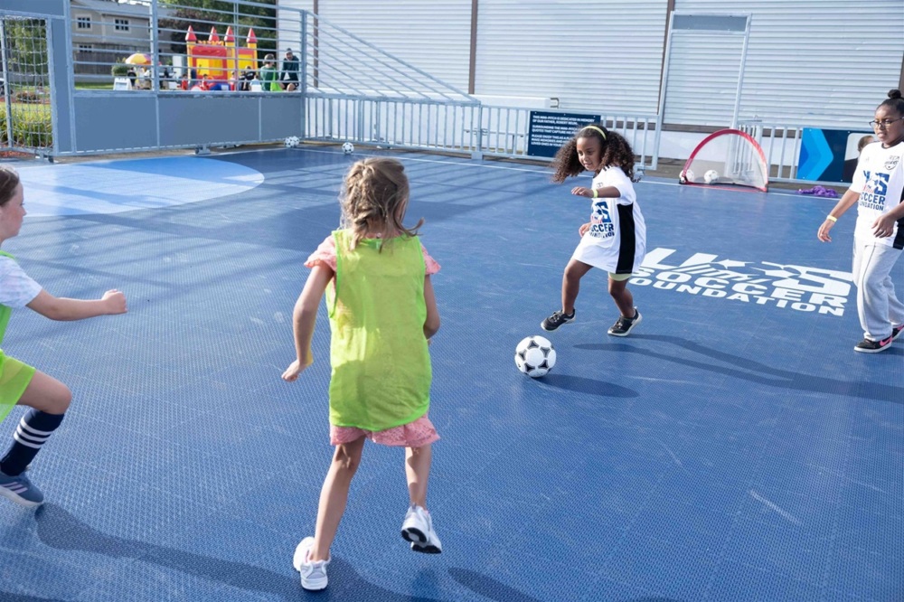 a young black looks down at a soccer ball getting ready to shoot against her opponents on the dark blue mini-pitch