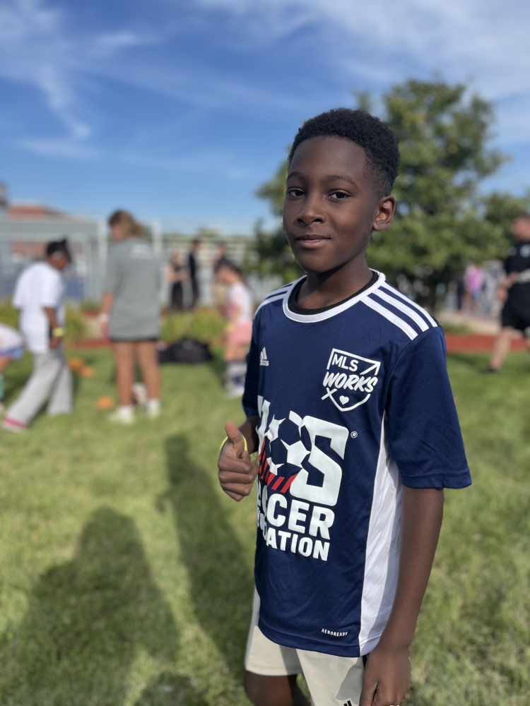 a young black boy gives a thumbs-up to the camera. he is wearing a navy blue U.S. Soccer Foundation jersey and a grin. He is on the grass.