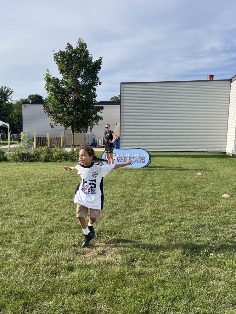 a young girl with long black hair celebrates scoring a goal in a fun jumping motion on the grass soccer pitch. she is wearing a white U.S. Soccer Foundation jersey.