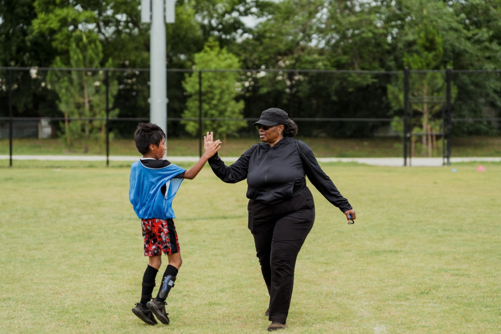 A coach-mentor dressed in black high-fives a young team member wearing a blue pinnie and patterned shorts on a grassy outdoor soccer field. The boy is mid-jump, and trees and a black fence are visible in the background, emphasizing their positive connection.