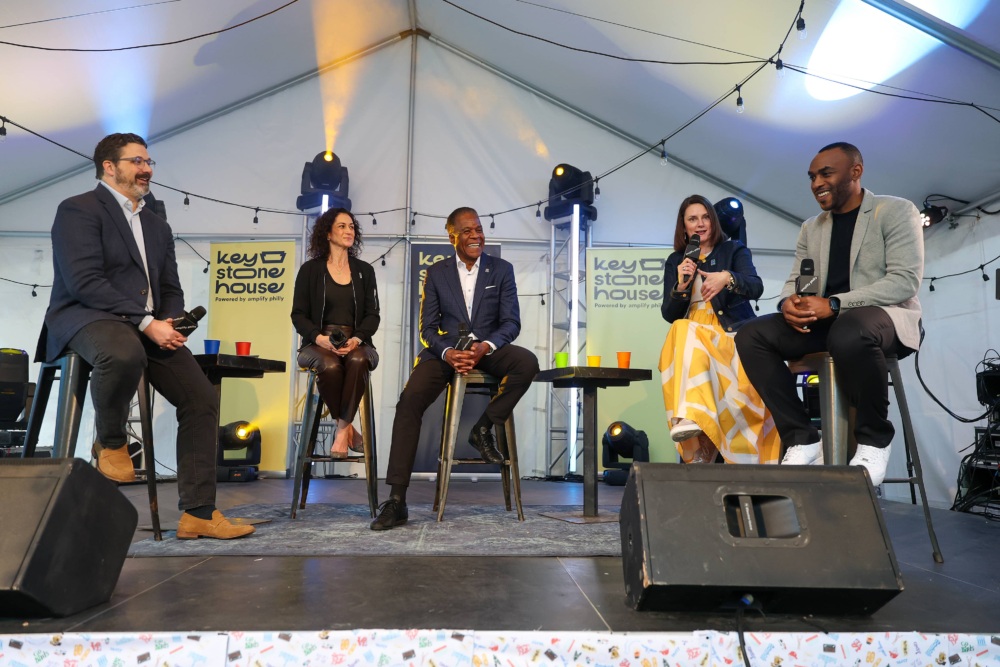 Five speakers sit on stools under a white event tent during the Yes, Coach!™ Initiative announcement at SXSW. They’re having a lively discussion, with branded Keystone House signs behind them and colorful lights adding to the atmosphere. The event highlights efforts to train coaches as mentors for youth.