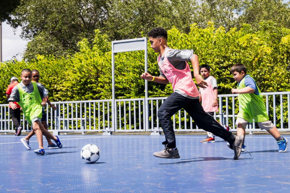 Boy in gray US Soccer Foundation t-shirt, pink pinni, and black pants chases a soccer ball as opposing team in green pinnies, runs in the background