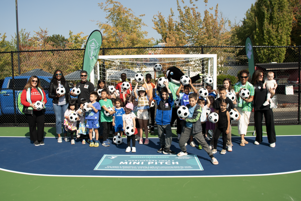 A group of adults and children pose with soccer balls on the Northshore YMCA mini-pitch opening