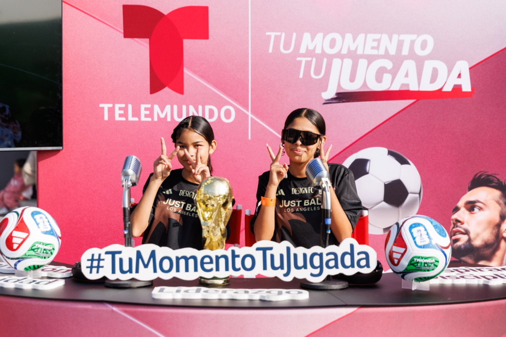 Two girls hold up peace signs with both hands in front of pink Telemundo #tuMomentoTuJugada banner