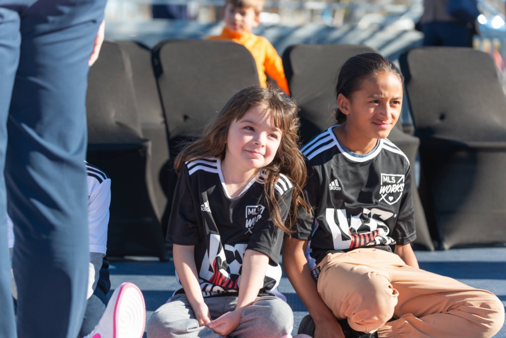 Two girls wearing U.S. Soccer Foundation jerseys sit on the outskirts of the mini-pitch, watching a game.