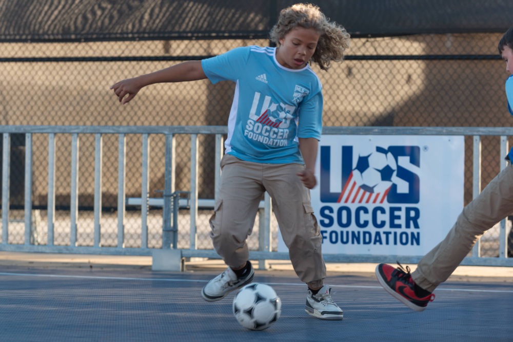 Kid wearing a light blue U.S. Soccer Foundation jersey kicks the soccer ball away from another player