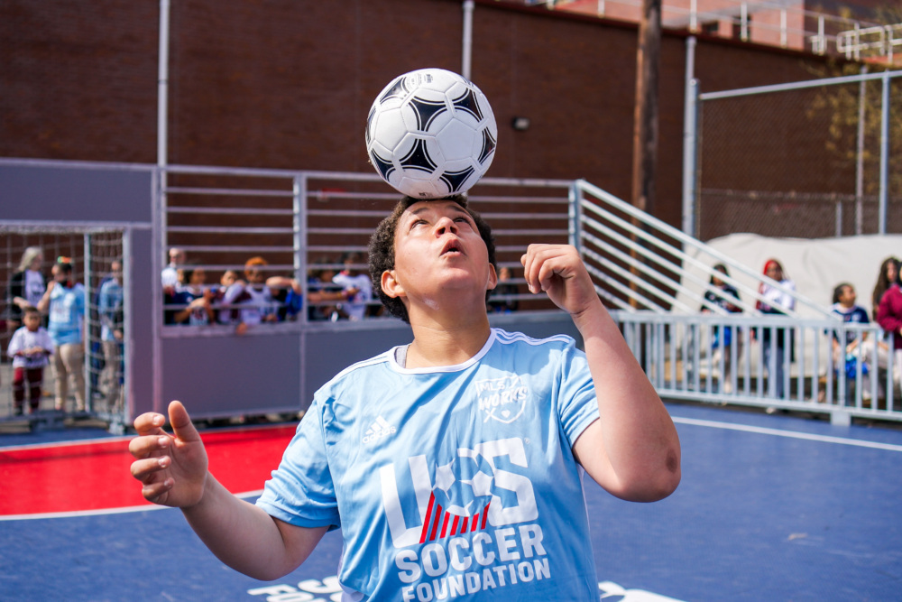Boy in U.S. Soccer Foundation jersey balances a soccer ball on his forehead,